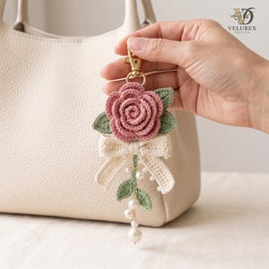 May include: A crochet keychain featuring a dusty rose flower with sage green leaves, a cream-colored bow, and a dangling gold-tone lobster clasp. The design includes small white faux pearls attached to the stem and a larger pearl at the bottom.