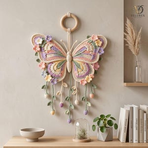 May include: A crochet butterfly wall hanging featuring pastel pink, yellow, purple, and green yarn. The butterfly is suspended from a wooden ring and decorated with small crocheted flowers and dangling vine-like strands with tiny buds.