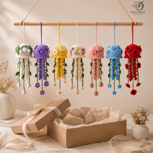 May include: Seven crocheted jellyfish ornaments in white, purple, yellow, cream, pink, blue, and red, each decorated with matching floral accents and hanging from a wooden rod. The ornaments feature dangling tentacles with small crocheted flowers.