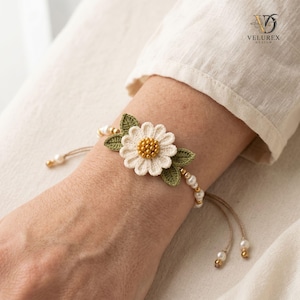 May include: A bracelet featuring a central crocheted white daisy with a golden beaded center, flanked by two green crocheted leaves. The band consists of small white pearls and gold beads on a thin cord, with dangling pearl and gold bead accents.