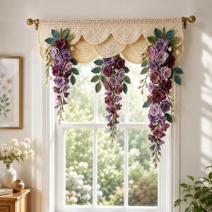 May include: A cream-colored crocheted window valance featuring a scalloped edge and three hanging floral arrangements. Each arrangement consists of cascading purple and burgundy crocheted flowers with green leaves, suspended from a brass curtain rod.