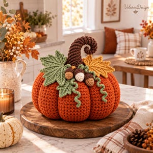May include: A crocheted orange pumpkin featuring a dark brown curled stem, a green maple leaf, a yellow maple leaf, and three small acorns. The pumpkin is decorated with green curly tendrils and sits on a round wooden board.