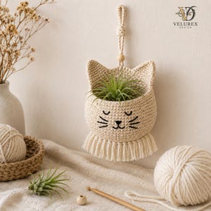 May include: A beige crocheted cat-shaped wall hanging planter featuring a small air plant inside, a hanging loop with a wooden bead, and a fringe trim at the bottom. The cat face has embroidered black eyes, a nose, and whiskers. VELUREX DESIGN text.