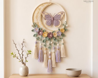 May include: A wall hanging featuring a crocheted crescent moon and a central lavender butterfly. The design includes crocheted flowers in yellow and purple, green leaves, and hanging tassels in cream and lavender. The text VELUREX DESIGN is visible.