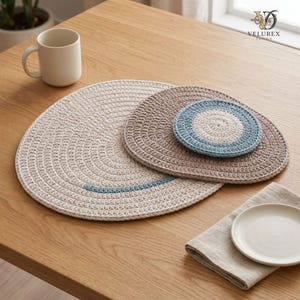 May include: A set of three round crocheted mats in cream, beige, and blue, featuring a large cream mat with a blue arc, a medium beige mat, and a small blue and cream coaster. A white ceramic mug and a small white plate on a linen napkin are nearby.