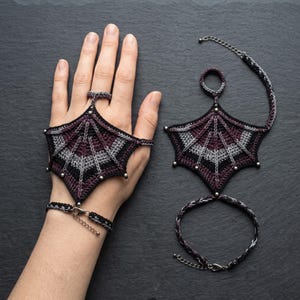 May include: A pair of crochet hand ornaments featuring a bat wing design in black, dark purple, and grey yarn. Each piece includes a ring loop, a central wing-shaped panel with metallic bead accents, and a wrist strap with a metal clasp closure.