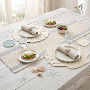 Peut inclure: Un service de table comprenant un chemin de table beige à rayures bleues, deux sets de table crème en forme de coquillage, deux assiettes blanches avec des serviettes en lin roulées et des ronds de serviette en corde, deux verres et des baies.