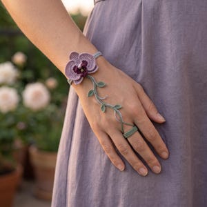Pode incluir: Uma pulseira de mão em crochê com uma grande flor de orquídea em tons de lavanda e roxo escuro, presa a uma faixa verde em forma de trepadeira que se estende pelo dorso da mão, conectando-se a um anel de crochê verde correspondente no dedo médio.
