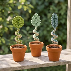 May include: Three small terracotta pots each containing a single crocheted plant stem. The stems feature spiral-wrapped yarn in shades of green and cream, topped with distinct crocheted leaf shapes in circular, serrated, and elongated designs.
