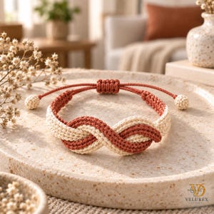 Könnte beinhalten: Ein geflochtenes Kordelarmband mit einem Unendlichkeitsknoten-Design in abwechselnden Terrakotta- und Cremetönen. Das Armband verfügt über einen verstellbaren Schiebeknotenverschluss mit zwei kleinen cremefarbenen Kugeln an den Schnurenden.
