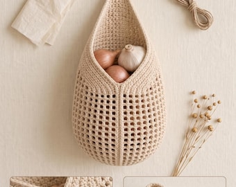 May include: A beige crocheted hanging basket with a grid-like pattern, featuring a top loop for suspension. The basket contains two small onions and a head of garlic. Included are a folded piece of fabric, a coil of twine, and a dried floral stem.