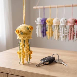 May include: A yellow crocheted jellyfish ornament featuring small yellow flower appliqués and green vine accents. The jellyfish has a long hanging cord and is displayed alongside a set of metal car keys on a light wood surface.
