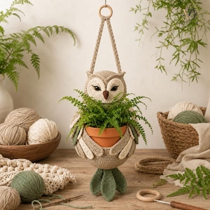 Peut inclure: Une jardinière suspendue en crochet en forme de hibou dans des tons beige, crème et vert sauge. Le hibou présente de grands yeux, un bec marron et des ailes qui soutiennent un pot en terre cuite contenant une fougère verte.