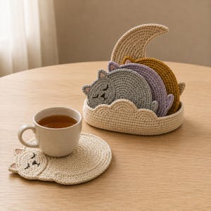 Puede incluir: Un juego de posavasos de ganchillo con forma de gato en gris, morado y amarillo mostaza, guardados en un soporte color crema con forma de nube y una luna creciente. Un posavasos de gato color crema está separado con una taza de té.