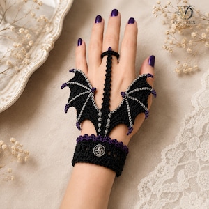 Puede incluir: Un accesorio de mano tejido en crochet negro con motivos de alas de dragón, detalles en violeta y cadenas plateadas. La pieza incluye una muñequera con un botón metálico circular y una anilla para el dedo, formando un arnés de mano.