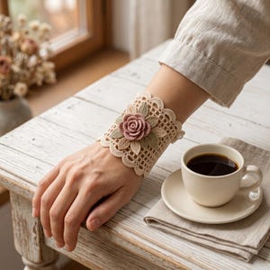 Peut inclure: Un bracelet de poignet en crochet beige orné d'une rose rose poudré centrale et de feuilles vert sauge. Le bracelet présente un motif en grille ajouré complexe et une bordure festonnée, offrant une texture détaillée et travaillée.