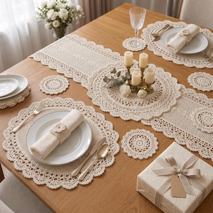 Puede incluir: Un camino de mesa, manteles individuales redondos y posavasos circulares de ganchillo color crema sobre una mesa de madera. El conjunto incluye servilleteros de ganchillo a juego con servilletas de tela, platos blancos y cubiertos dorados.