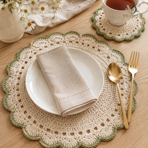 Op de afbeelding: Een ronde, crèmekleurige gehaakte placemat met een geschulpte saliegroene rand, voorzien van een wit keramieken bord met een gevouwen beige linnen servet, een gouden lepel, een gouden vork en een bijpassende gehaakte onderzetter met theekop.
