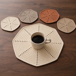 May include: A set of five octagonal crochet coasters in cream, beige, and terracotta colors. One large cream coaster supports a ceramic mug filled with black coffee, while four smaller coasters are arranged around it on a wooden surface.