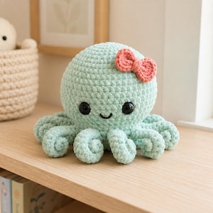 May include: A light mint green crocheted octopus plush toy with a round head and eight curled tentacles. The octopus features two large black plastic safety eyes, a small embroidered black smile, and a small coral pink crocheted bow.