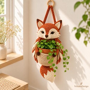 Puede incluir: Un macetero de pared tejido a ganchillo con forma de zorro, hecho de hilo color naranja óxido y crema, con un bolsillo central que sostiene una maceta de terracota con una planta verde colgante. El zorro tiene ojos oscuros y una cola larga.