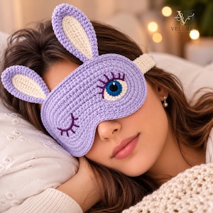 May include: A lavender crochet sleep mask featuring two upright bunny ears with cream-coloured centres. The mask displays a winking eye with dark purple eyelashes on the left and an open blue and white eye with dark purple eyelashes on the right.