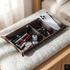 May include: A dark brown leather valet tray with multiple compartments containing a white smartphone, a black car key fob, a silver wristwatch with a red and blue bezel, several pens, and a black braided bracelet with silver accents.