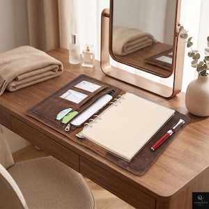May include: A brown leather portfolio organizer featuring a six-ring binder mechanism with lined paper, multiple card slots, a zippered compartment, and a pen loop holding a red and silver pen, all laid open on a wooden surface.