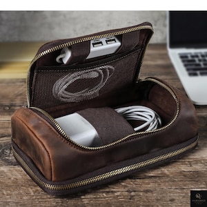 May include: A brown leather zippered travel organizer case with a structured cylindrical shape. The interior features a mesh pocket, elastic straps, and compartments holding a white USB hub and a coiled white charging cable for electronic devices.