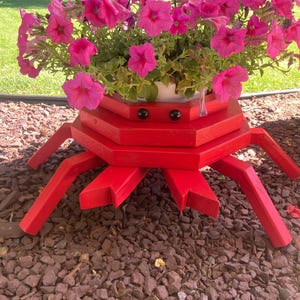 May include: A bright red wooden crab-shaped planter featuring a tiered octagonal body, two small black circular eyes, and eight angled legs. The planter holds a hanging basket filled with vibrant pink petunia flowers with green foliage.
