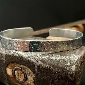 Peut inclure: Un bracelet manchette en métal argenté présentant un motif texturé répétitif sur toute sa surface extérieure. Le bracelet possède un design ouvert avec des extrémités arrondies, arborant une finition polie qui reflète la lumière.