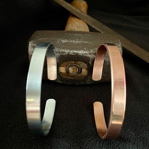 May include: Two open cuff bracelets, one in a silver-toned metal and one in a copper-toned metal, both featuring a smooth, flat surface. The bracelets are positioned in front of a heavy metal hammer with a wooden handle and a metal file.