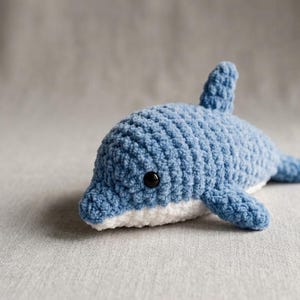 Può includere: Un piccolo delfino all'uncinetto fatto a mano, realizzato con filato blu morbido sulla parte superiore e bianco sul ventre. Il peluche presenta una pinna dorsale prominente, due pinne laterali, una coda e un occhio di sicurezza nero visibile.