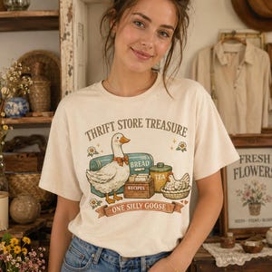 May include: A cream-colored short-sleeve t-shirt featuring a graphic print of a goose wearing a bow tie, a bread box, a tea canister, and a basket with a chicken. The text reads THRIFT STORE TREASURE and ONE SILLY GOOSE in brown lettering.