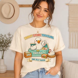May include: A cream-colored t-shirt featuring a graphic of a white goose inside a teal bread box, a small chicken in a basket, and boxes labeled Recipe and Tea. The text reads THRIFT STORE TREASURE and ONE SILLY GOOSE in colorful lettering.
