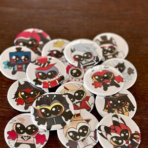 May include: A collection of circular pin-back buttons featuring various cartoon characters with large black eyes. Each button displays a unique character design with distinct color schemes, including red, black, white, and blue, arranged in a scattered pile.