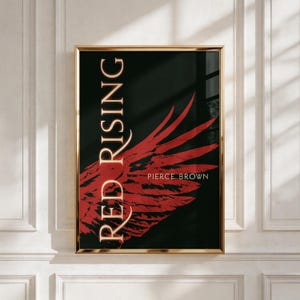 May include: A rectangular wall art print in a gold frame featuring a black background with a large red wing graphic. The text RED RISING is printed vertically in a cream serif font on the left, with PIERCE BROWN in smaller text on the right.