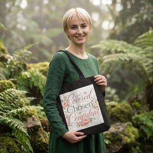 Può includere: Una borsa tote nera con un pannello rettangolare bianco riportante il testo Covered, Chosen, Confident in un carattere corsivo decorativo, circondato da un'illustrazione floreale di rose rosa e foglie verdi su uno sfondo bianco.
