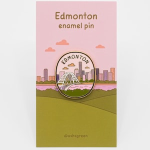 May include: A round enamel pin featuring an illustration of the Edmonton skyline and bridge, displayed on a rectangular card with the text Edmonton enamel pin and @ashsgreen. The pin shows a city silhouette, a white bridge, and a river.