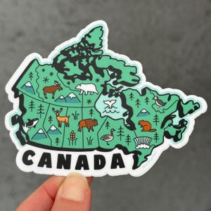May include: A sticker in the shape of Canada, featuring a green map with illustrations of wildlife and landmarks. The word "CANADA" is printed in bold white letters at the bottom. The sticker is held by a hand.