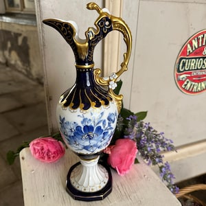 Op de afbeelding: Een porseleinen kan met een diepblauw en wit kleurenpalet en handgeschilderde blauwe bloemmotieven. De kan heeft sierlijke goudkleurige accenten op het handvat, de rand en de voet, met een geschulpte hals en een voetstuk.