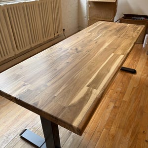 May include: A rectangular standing desk featuring a natural wood grain tabletop with visible light and dark brown tones. The desk is supported by a black metal frame with two vertical columns and flat, horizontal feet resting on a wooden floor.