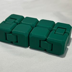 May include: A dark green infinity cube fidget toy composed of eight smaller connected blocks. The object features a smooth matte finish and is constructed with hinged joints that allow the blocks to fold and rotate into various geometric shapes.