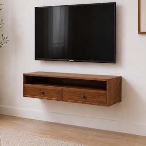 May include: A wall-mounted rectangular wooden media console featuring a dark brown finish, an open top shelf, and two lower drawers with small dark circular knobs. The console is positioned directly beneath a large black flat-screen television.