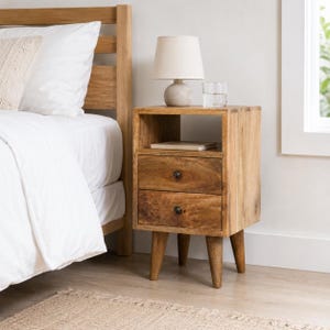 May include: A light brown wooden nightstand featuring an open storage shelf at the top, two drawers with small dark circular knobs, and four tapered legs. A beige lamp and a clear glass of water sit on the flat top surface of the nightstand.