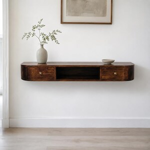 May include: A dark brown wooden floating wall shelf with rounded edges, featuring two drawers with small round brass knobs and a central open storage compartment. The shelf is mounted horizontally on a plain white wall, providing a sleek surface.