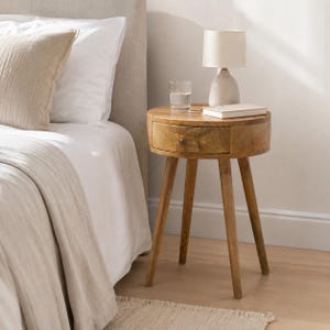 May include: A round wooden bedside table with a single drawer and four tapered legs. On top of the table sits a small white lamp, a clear glass of water, and a closed book. The table is positioned next to a bed with white bedding and a pillow.