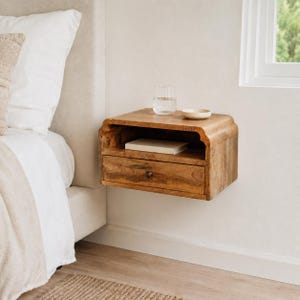 May include: A wall-mounted wooden nightstand featuring a natural light brown wood grain finish, a single storage drawer with a small dark knob, and an open shelf above the drawer. A clear glass and a small white bowl sit on the top surface.