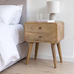 May include: A light brown wooden nightstand with rounded corners, two drawers featuring small round brass knobs, and four tapered legs. A cylindrical lamp with a white shade and a clear glass tumbler sit on top of the wooden surface.