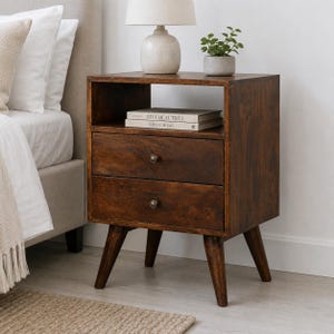 May include: A dark brown wooden nightstand with two drawers, each featuring a single round knob. The nightstand has an open storage shelf at the top and four tapered legs. Two books are stacked on the shelf, titled Live Beautiful and This Is Home.